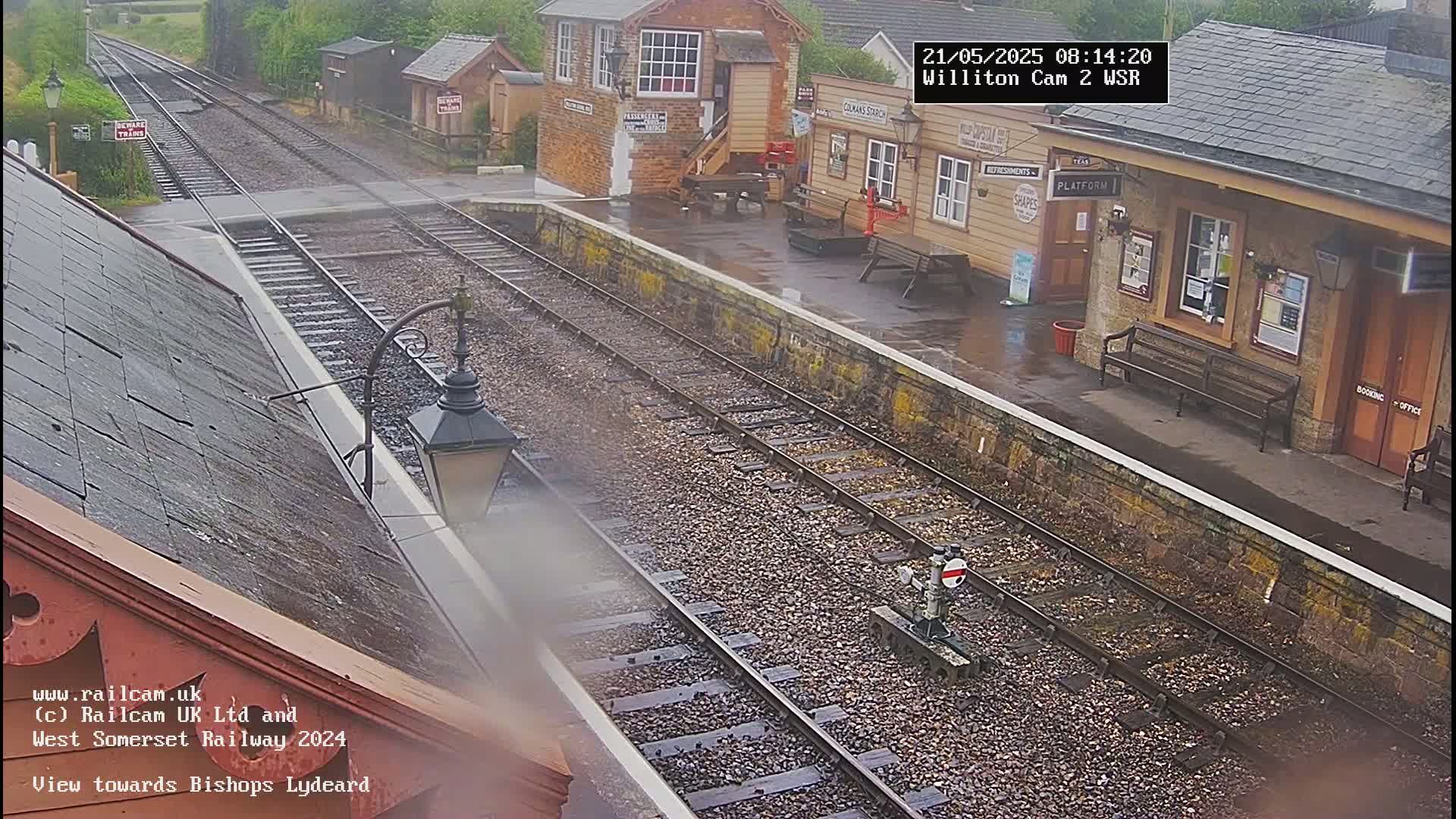 A wet train station platform with train tracks and buildings is viewed from a high vantage point in the rain.