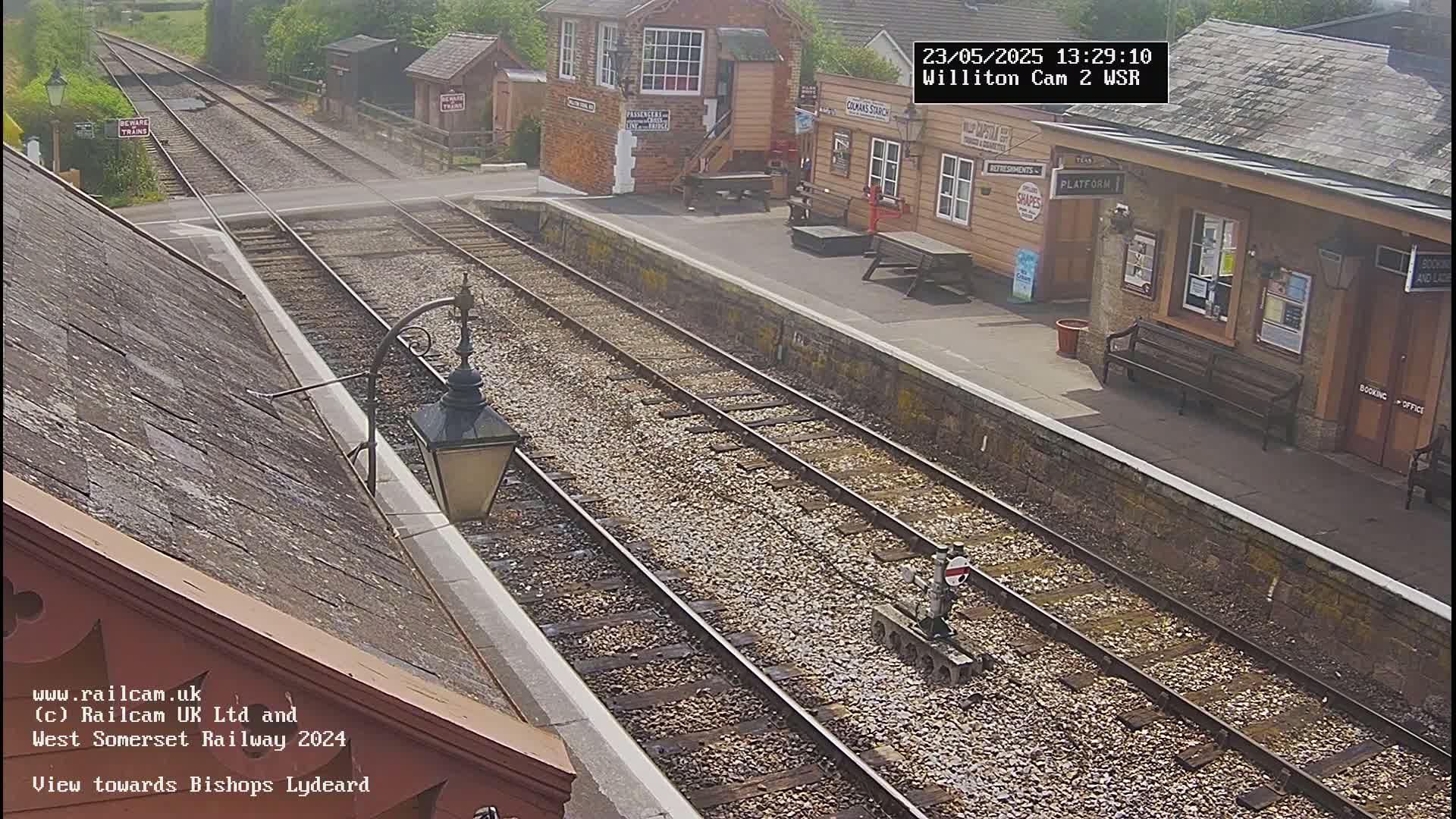 A train station with two sets of train tracks, a small train on one track, and several buildings on a sunny day.