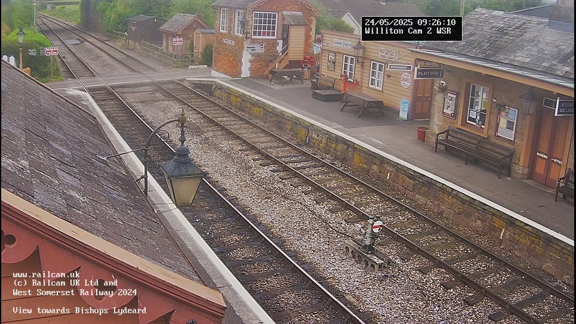 A view from a train station platform shows two sets of train tracks extending into the distance under an overcast sky, with a small station building and bench visible.