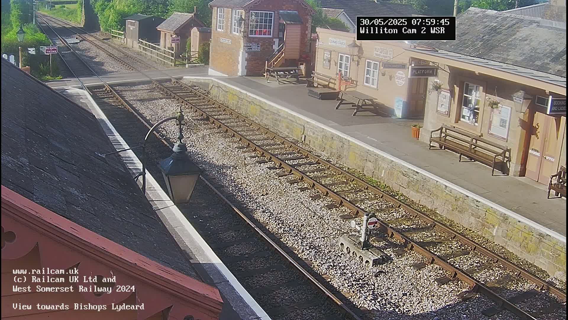 A sunny view of a railway station platform with train tracks extending into the distance, and a signal box and station building visible.