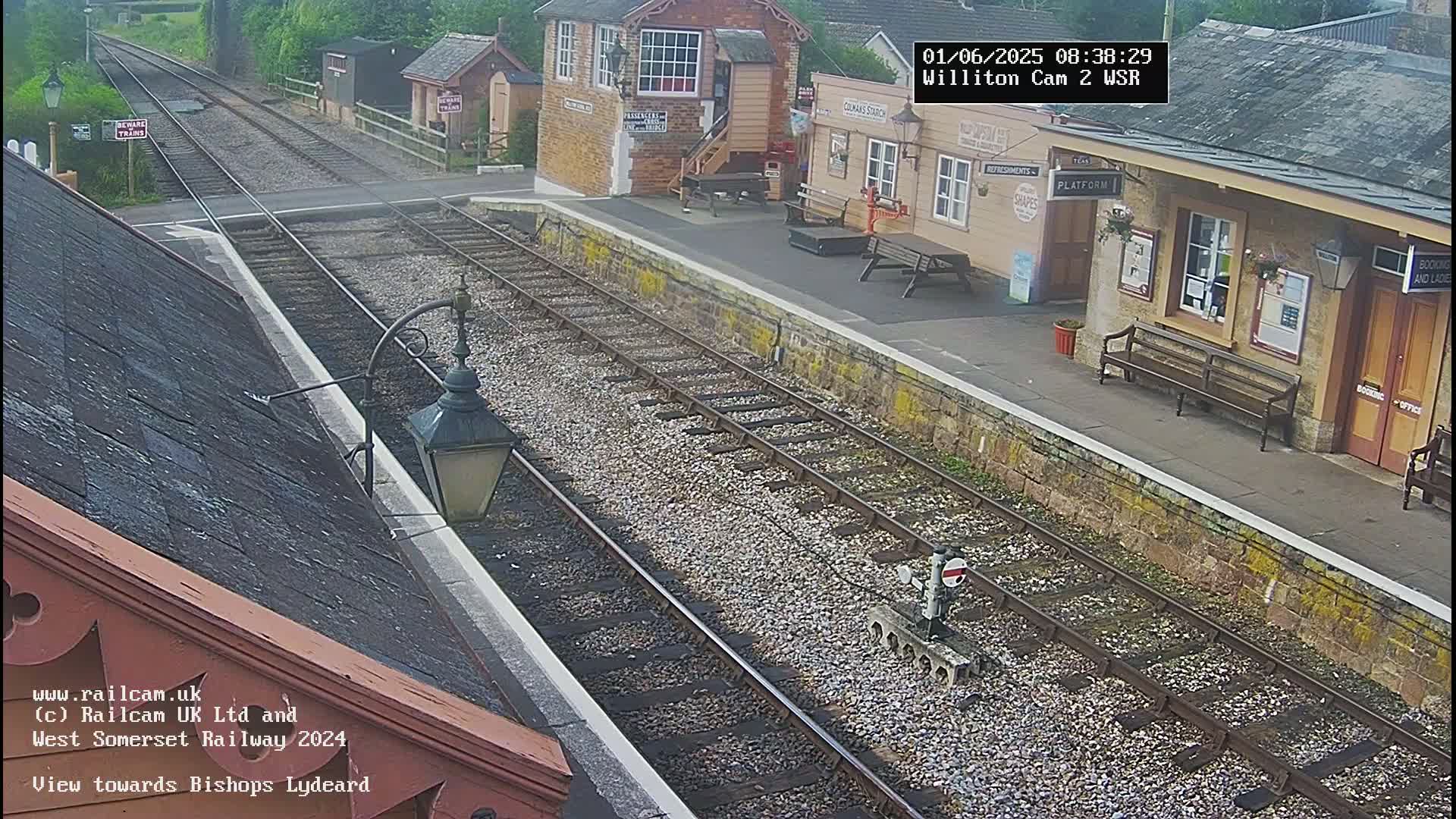 A high-angle view of a train station platform on a partly cloudy day, showing train tracks extending into the distance.