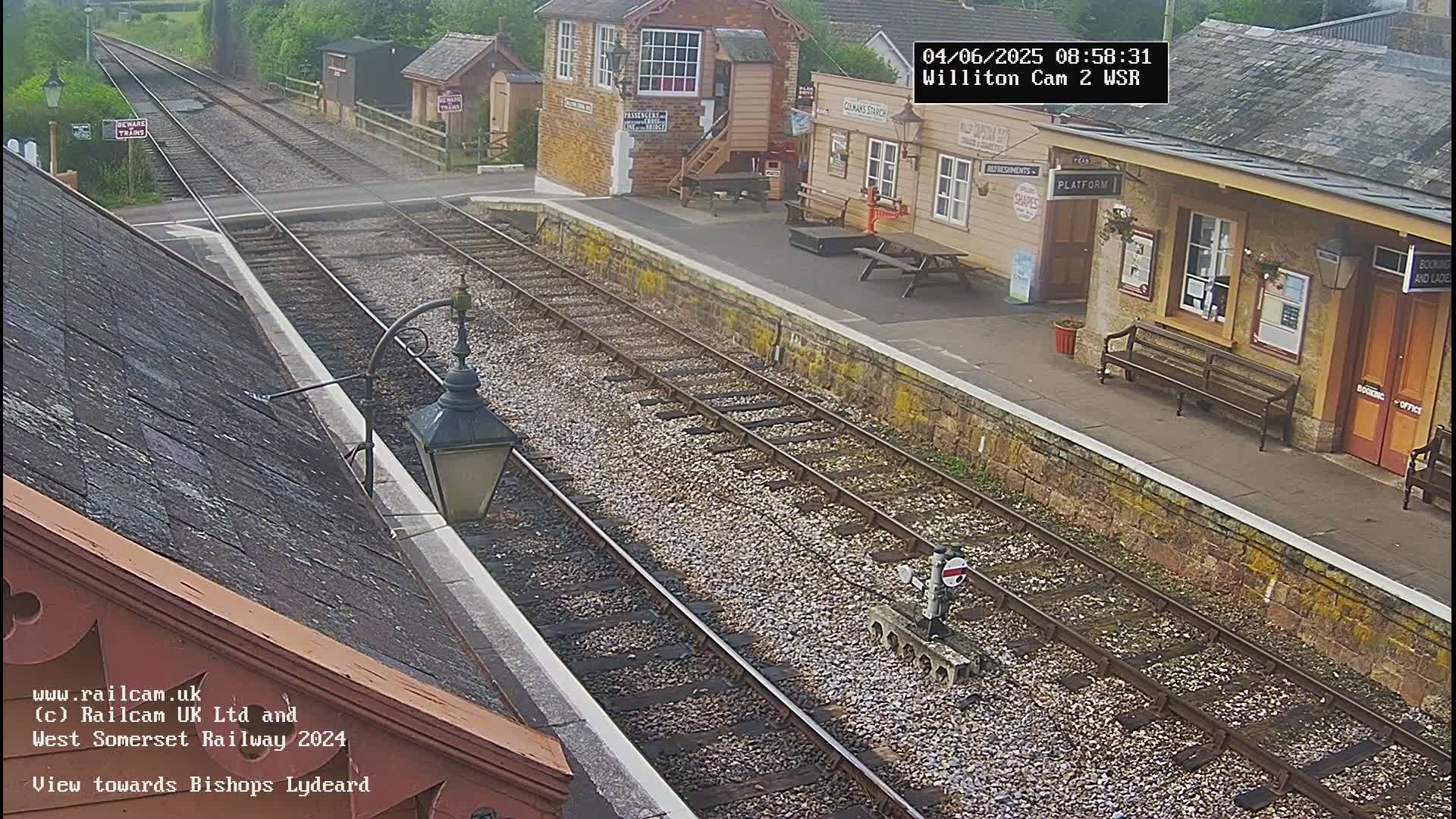 A train station platform with railway tracks extending into the distance under an overcast sky.