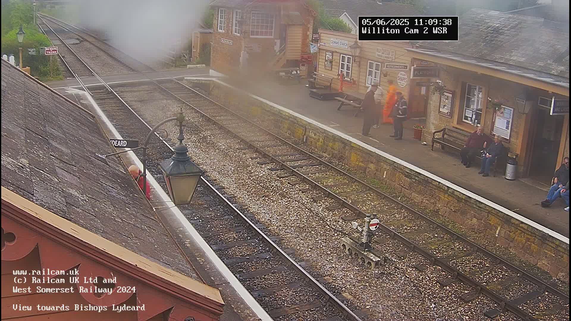 A hazy view of a train station platform with several people standing and sitting near a building, train tracks, and a partially visible steam plume.