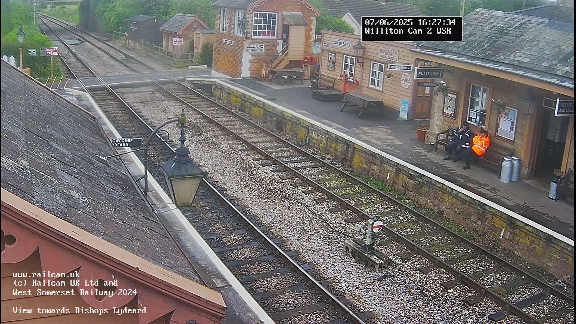 A high-angle view of a train station platform with two people sitting on a bench, railway tracks extending into the distance under an overcast sky.