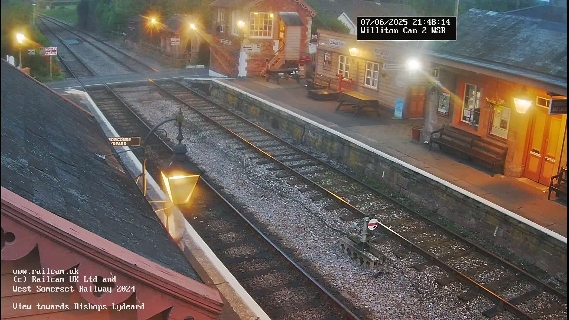 A nighttime view of a train station with two sets of train tracks, lit by streetlights and building lights.