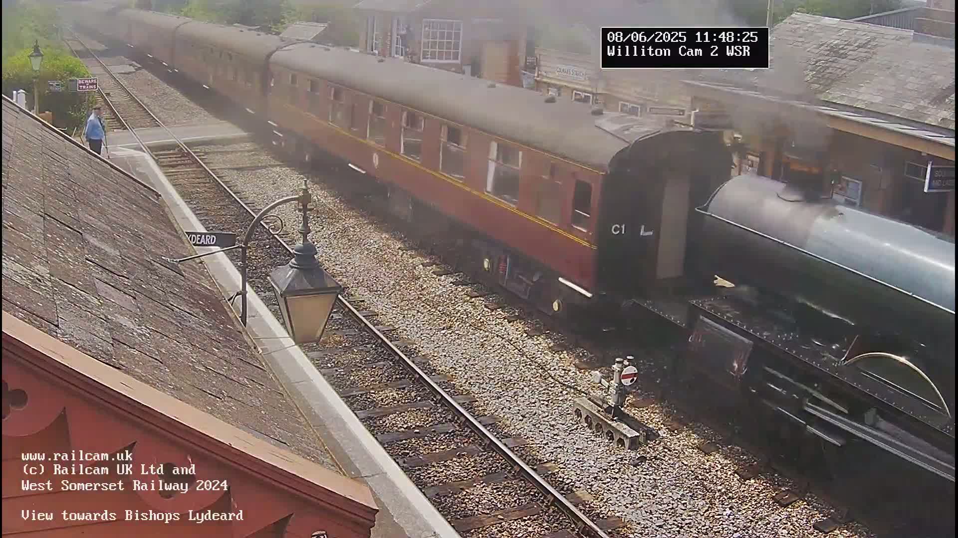 A steam train passes a station on a sunny day, with a person standing on the platform.