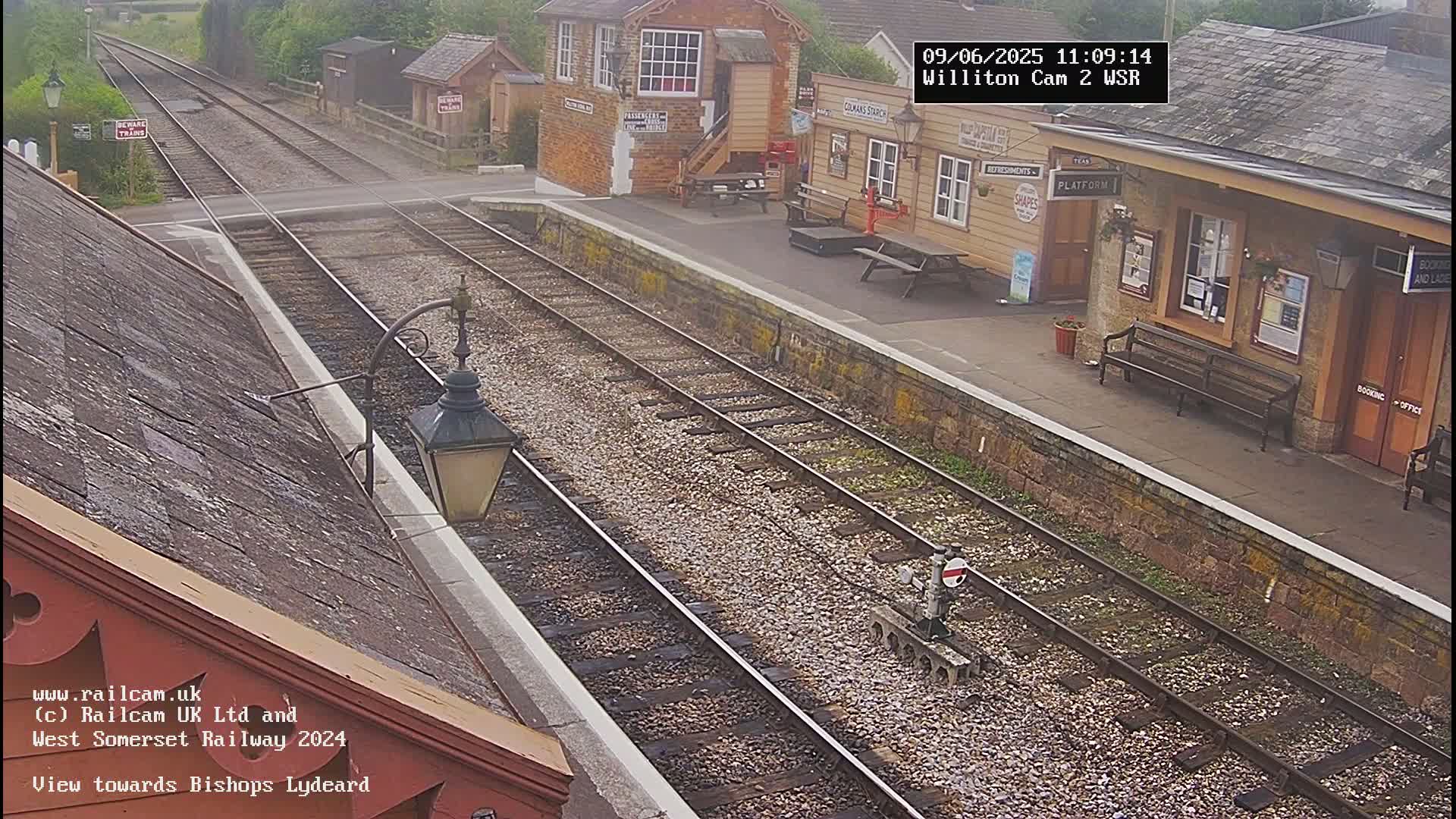 A train station platform with two parallel train tracks, a signal, and several buildings under an overcast sky.