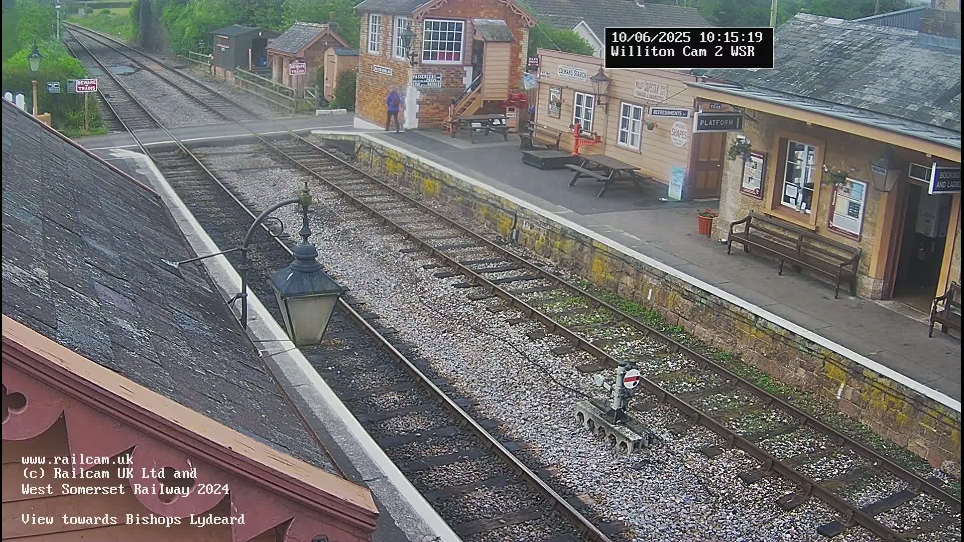 A train station platform with two sets of train tracks, a small building, and several benches is seen on an overcast day.