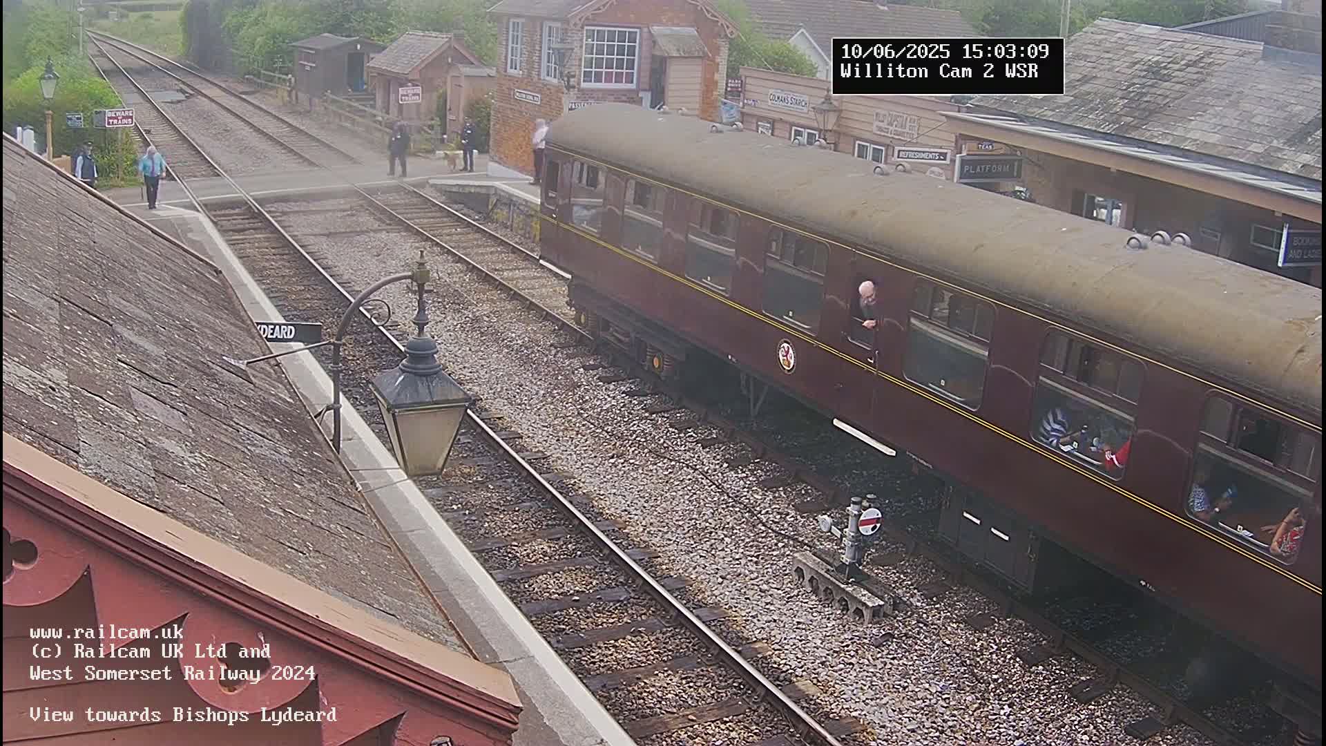 A maroon railway carriage is partially visible at a train station on an overcast day, with several people standing on the platform nearby.