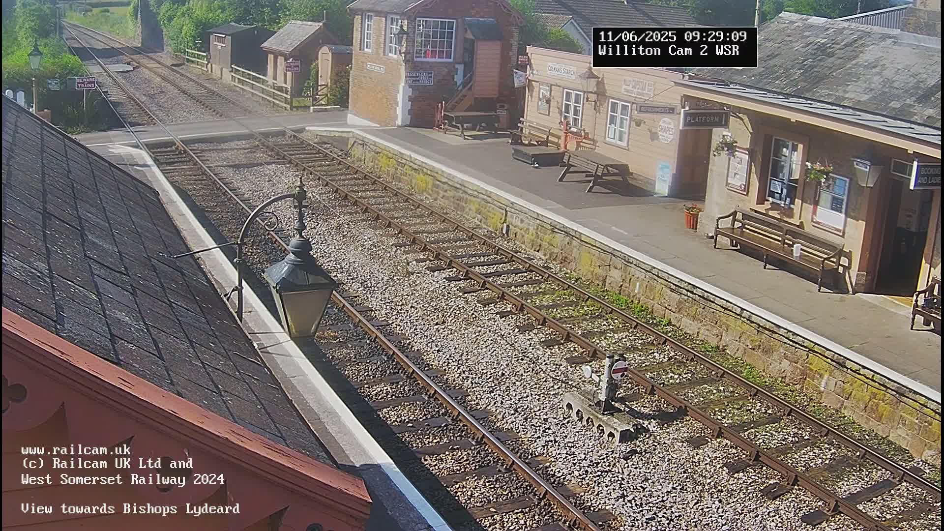 A sunny view of a train station platform with two sets of train tracks extending into the distance.