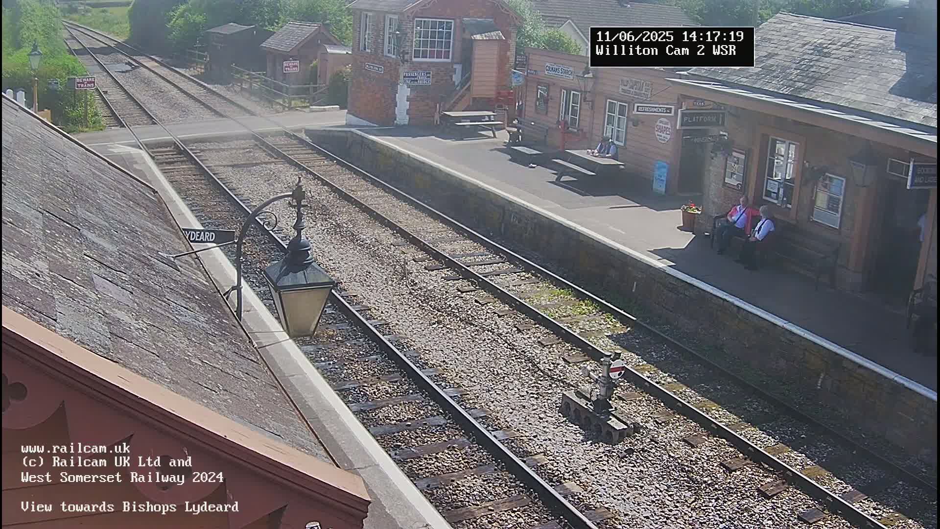 A sunny view of a train station with two people sitting on a bench, and train tracks extending into the distance.