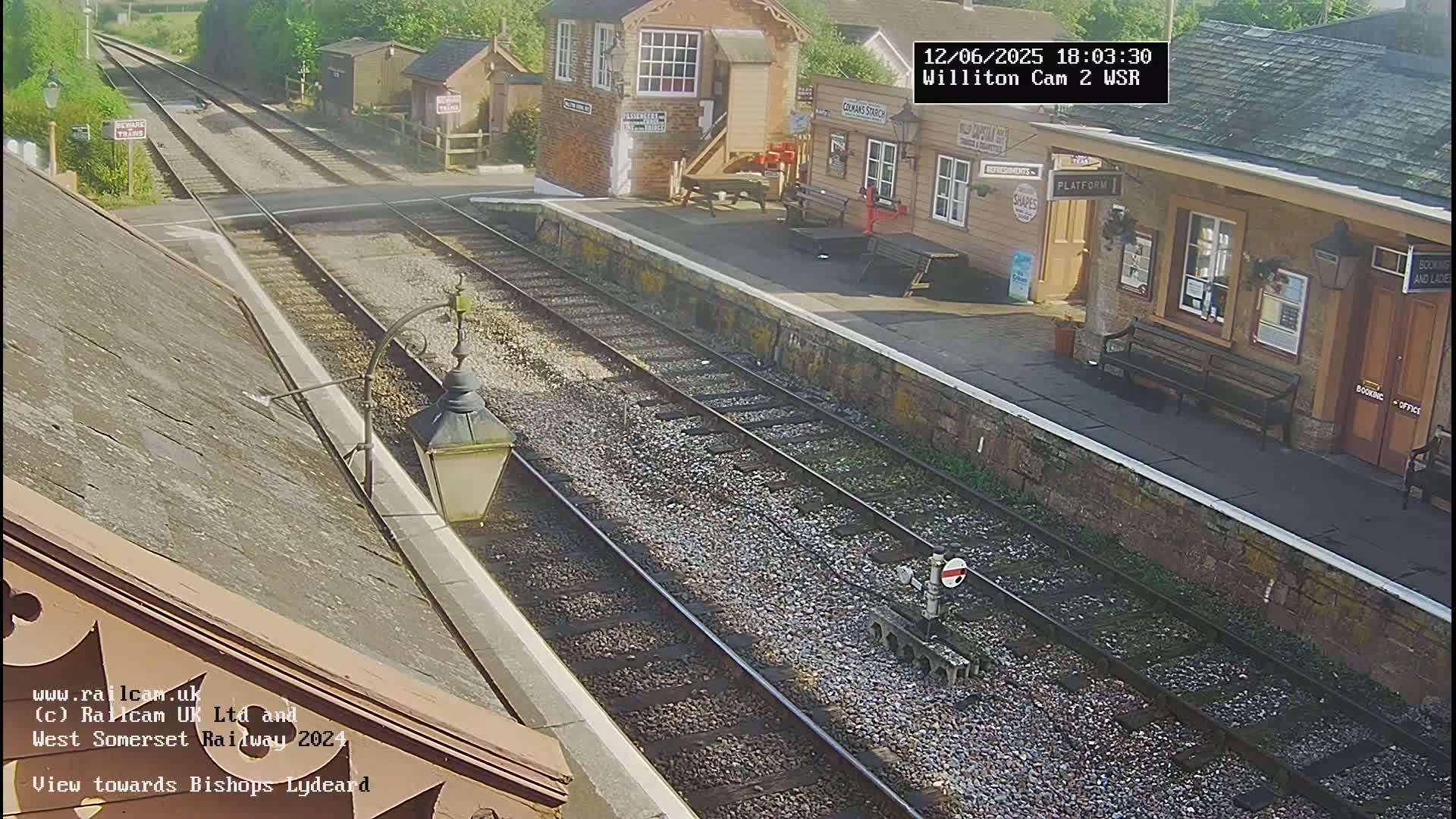 A sunny view of a train station with two sets of train tracks, a small building, and a bench.