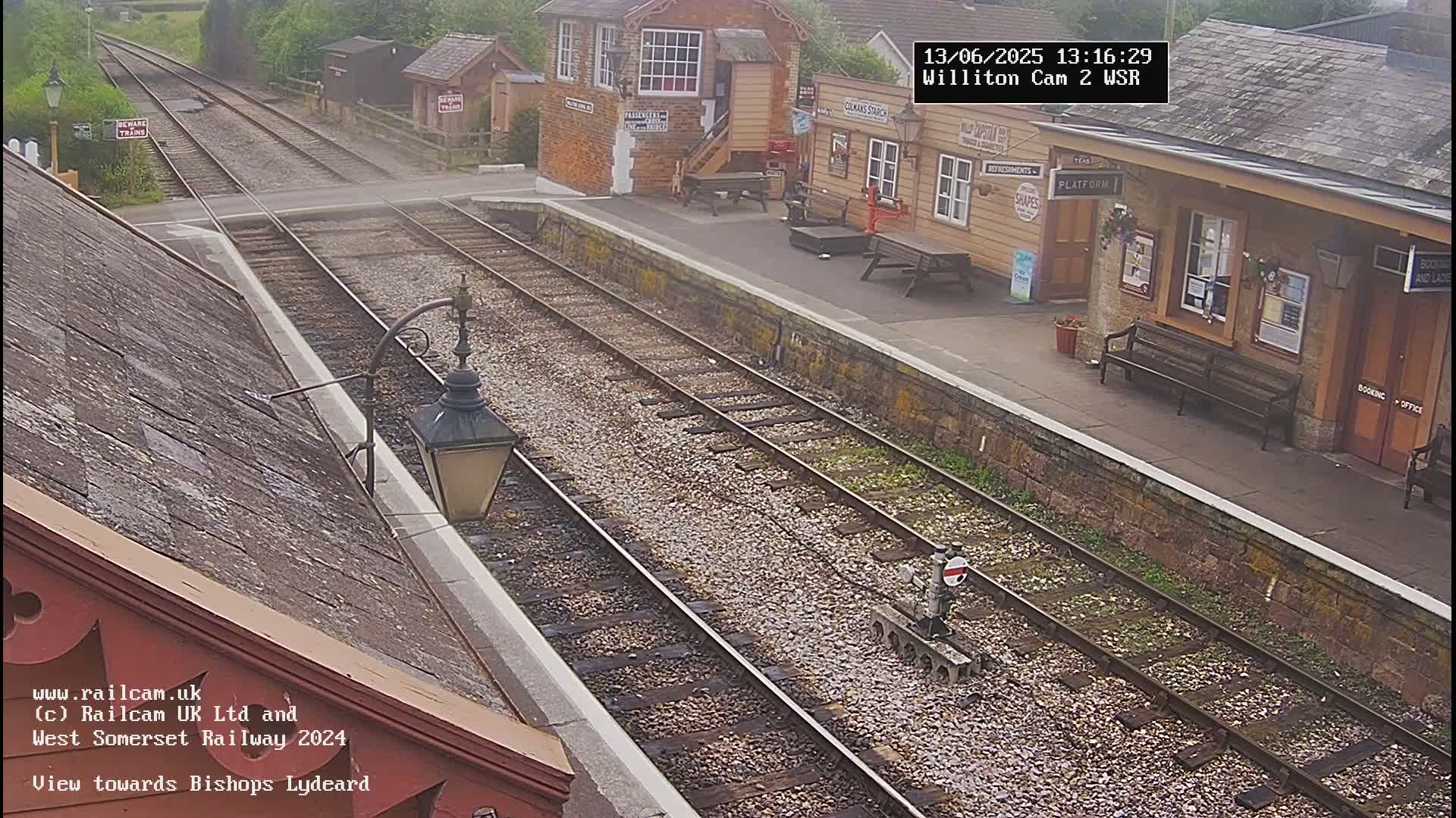 A high-angle view of a train station on an overcast day shows two parallel train tracks extending into the distance, with a small station building and platform visible.