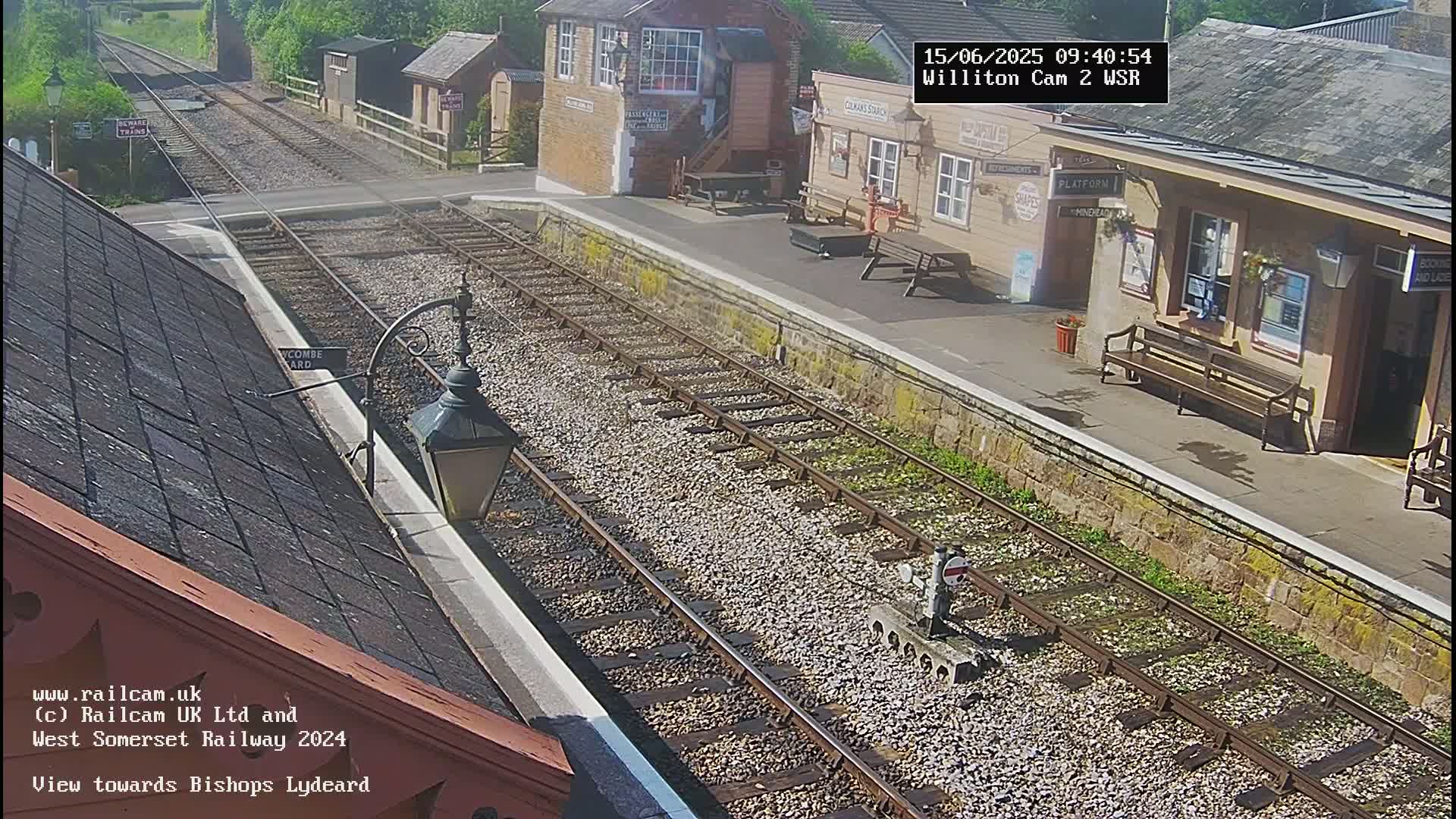 A sunny view of a train station platform with two parallel train tracks and a building.
