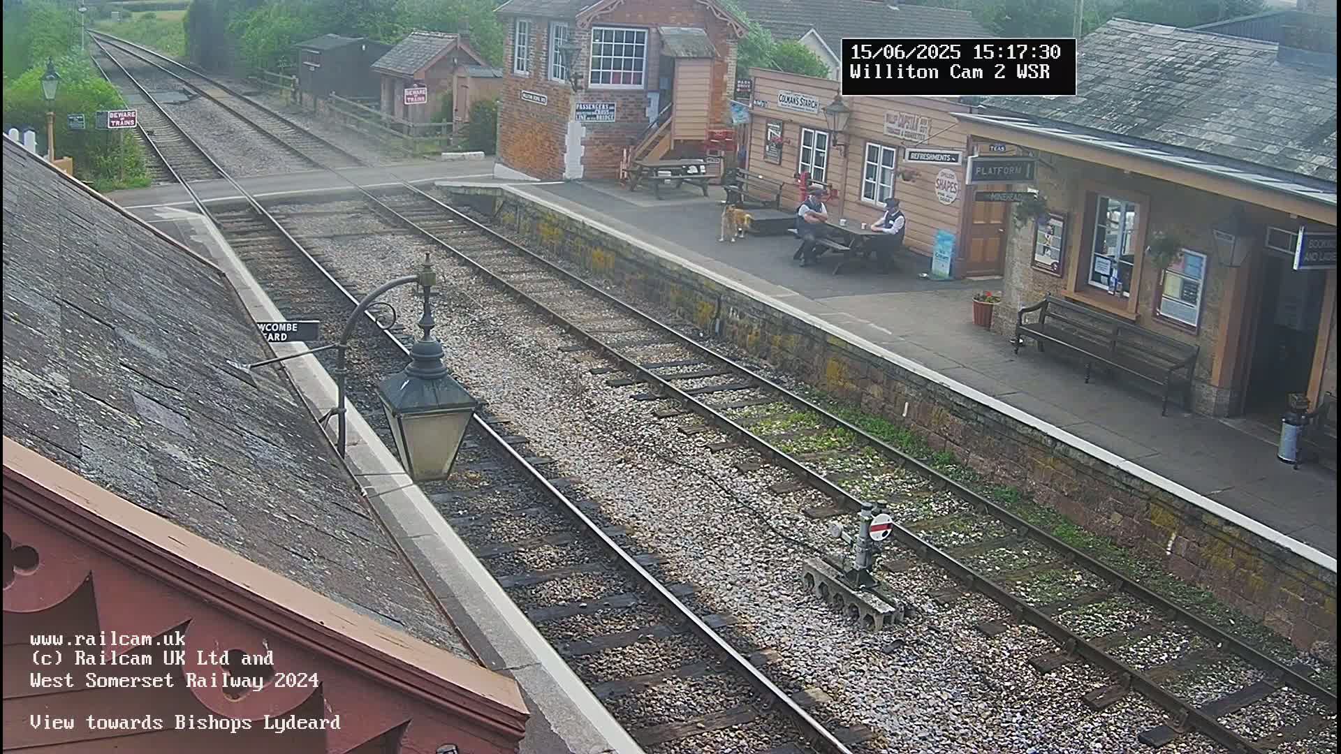 A high-angle view shows a train station platform with two people sitting on a bench, a dog nearby, and railway tracks extending into the distance under a partly cloudy sky.