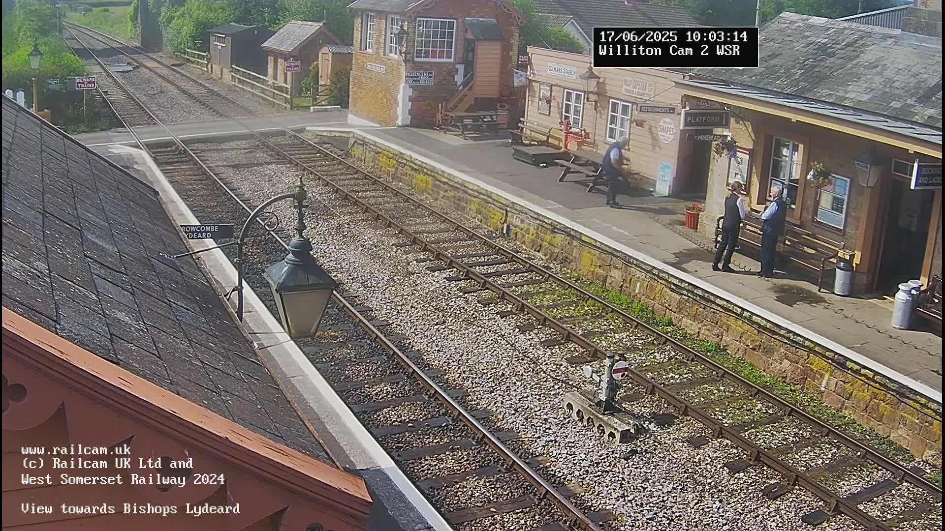 A sunny view of a train station platform with two sets of train tracks, several people standing near a building, and a lamppost on the left.