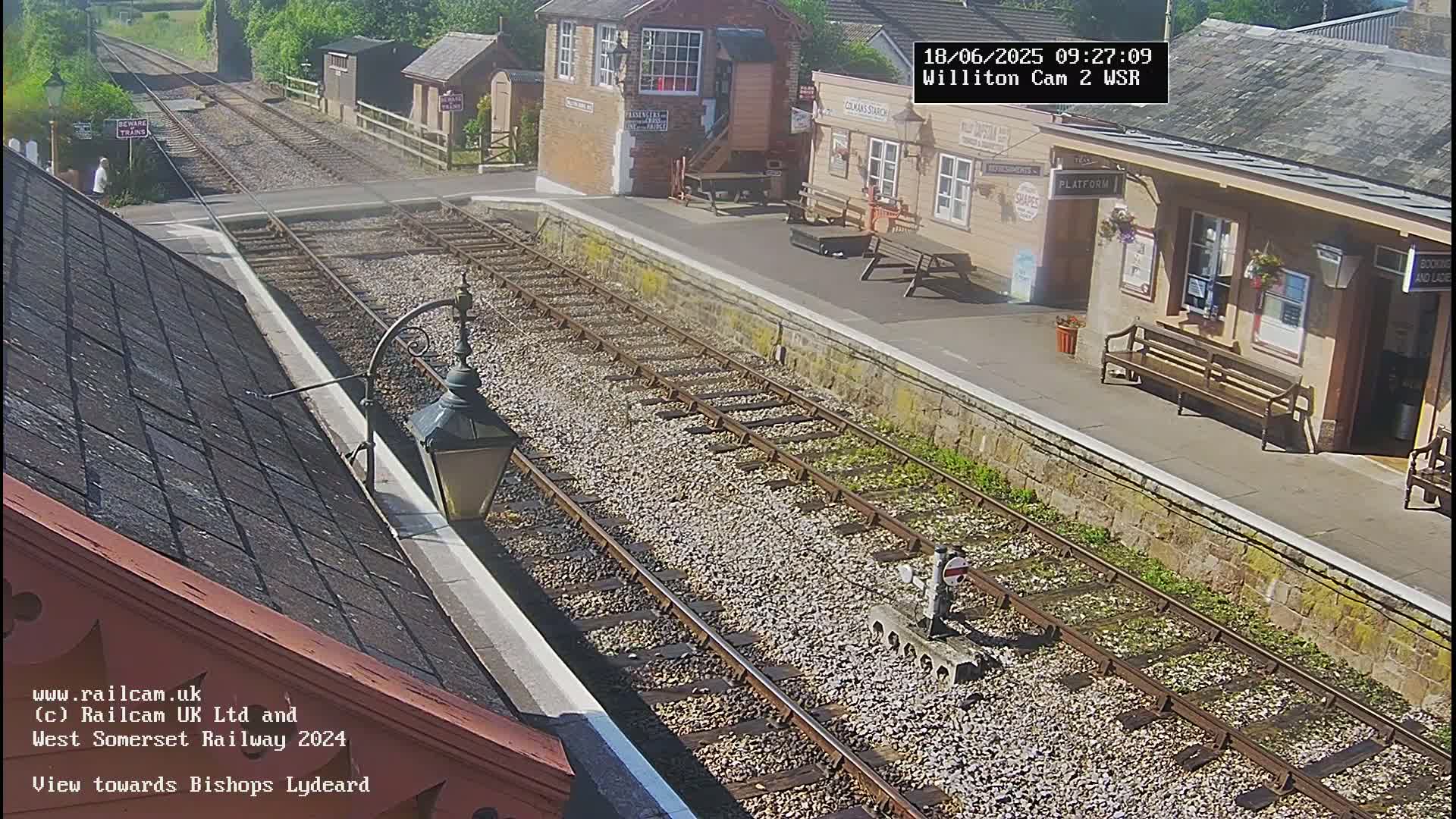 A sunny view of a train station platform with multiple train tracks, a signal box, and benches.