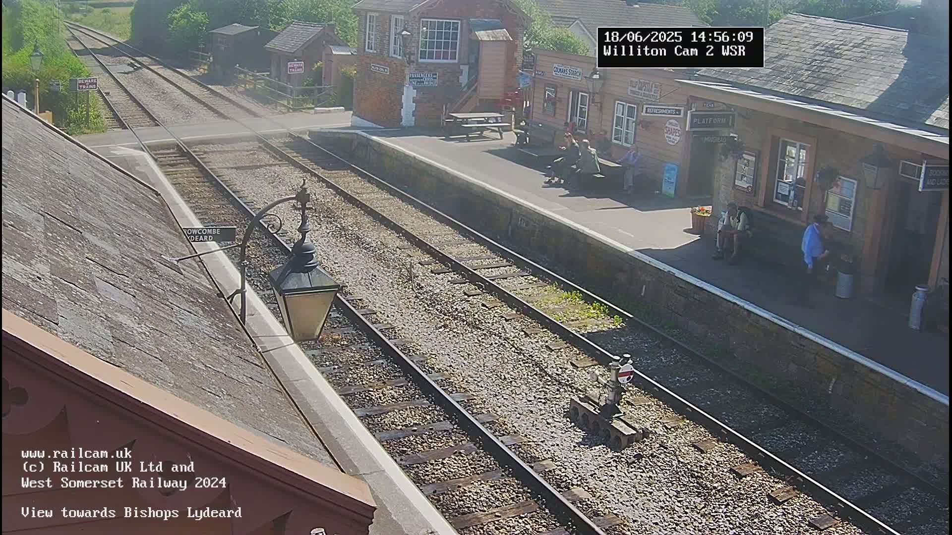 A sunny view of a train station platform with several people waiting, railway tracks extending into the distance, and buildings visible in the background.
