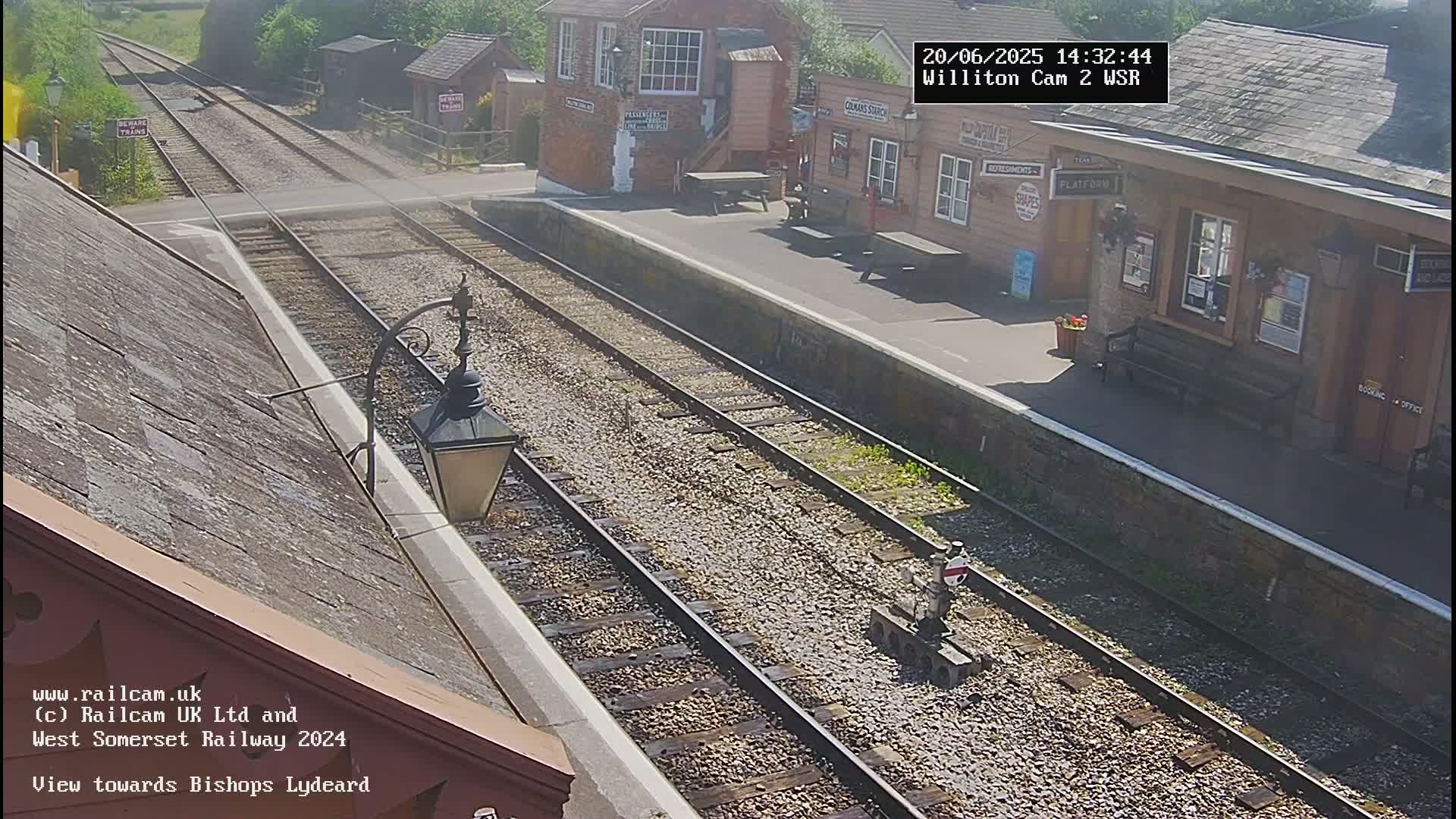 A sunny view of a train station platform with multiple train tracks, buildings, and a lamppost.