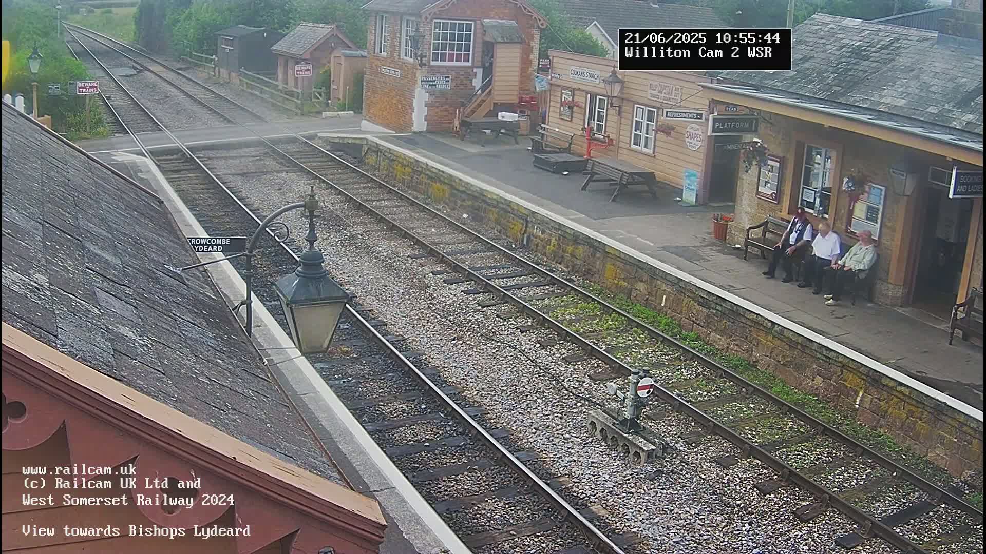 A train station platform with several people sitting on benches, viewed from above on an overcast day, shows two sets of train tracks leading away from the station.