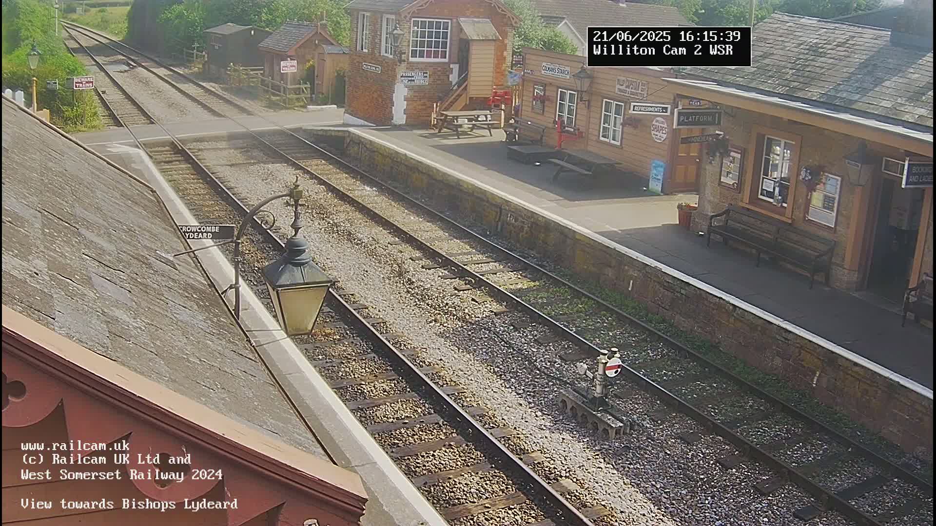 A sunny view of a train station with several train tracks and a platform, showing a wooden building and a bench.