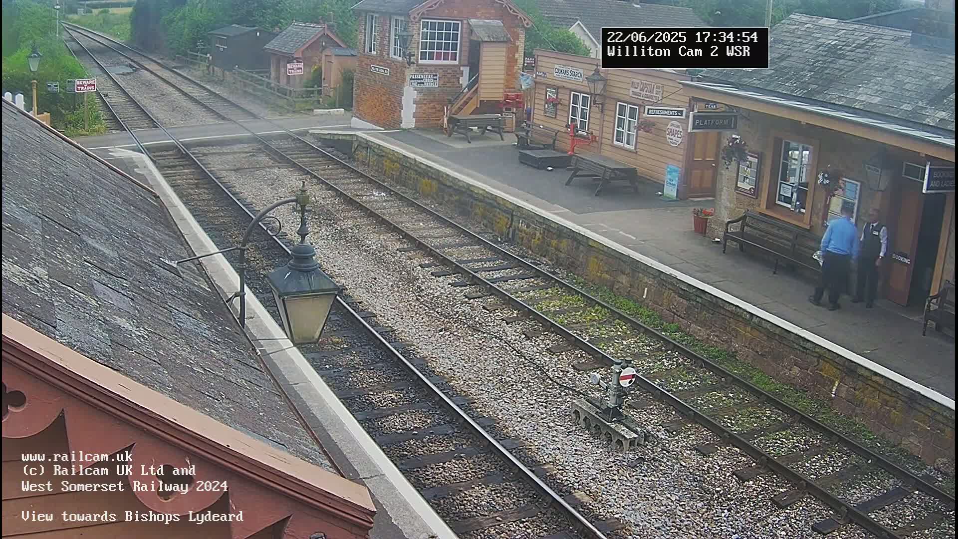 A railway station platform with two people standing near a building, and train tracks extending into the distance under an overcast sky.