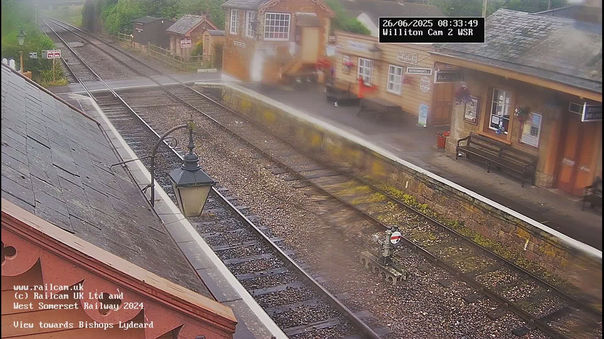 A train passes a railway station on a misty day, with a platform and buildings visible.
