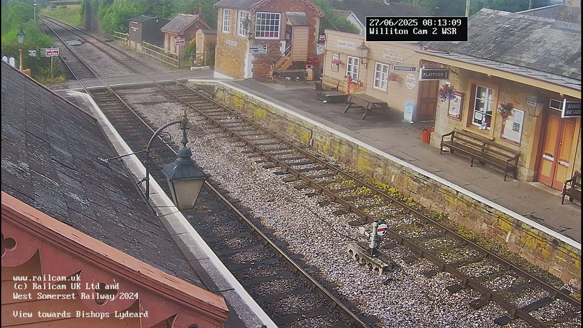 A train station platform with two sets of tracks, a stone building, and benches sits under an overcast sky.