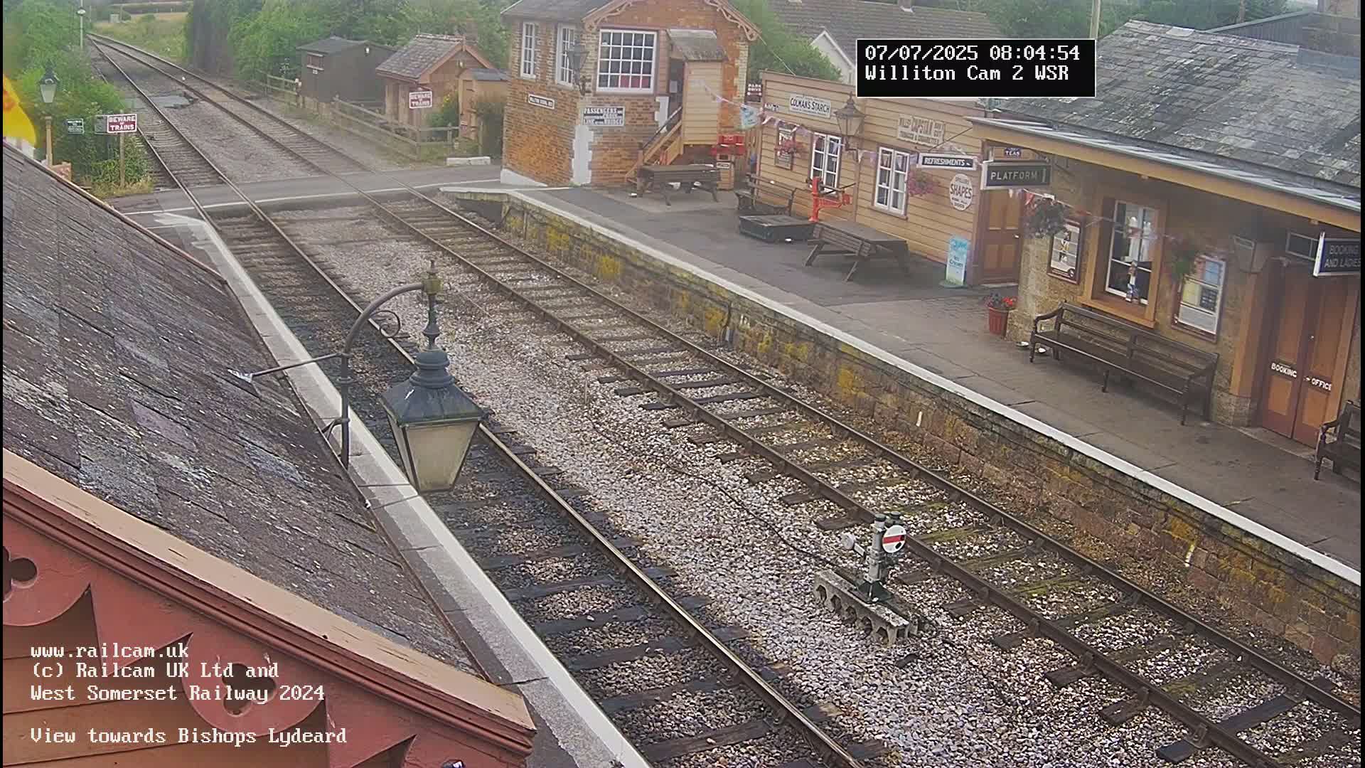 A train station platform with railway tracks, a signal, and several buildings, viewed on an overcast day.