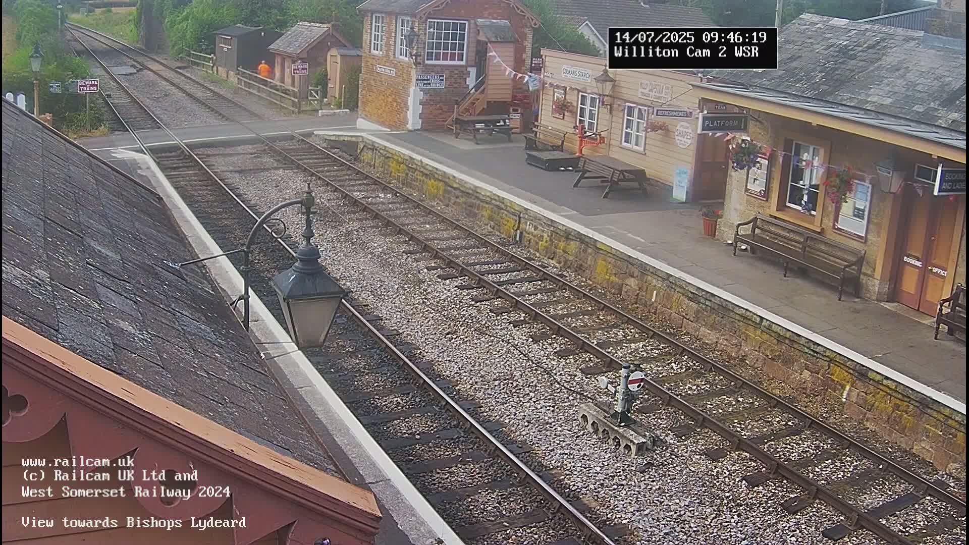 A train station platform with a signal box and a small waiting area, under a partly sunny sky.