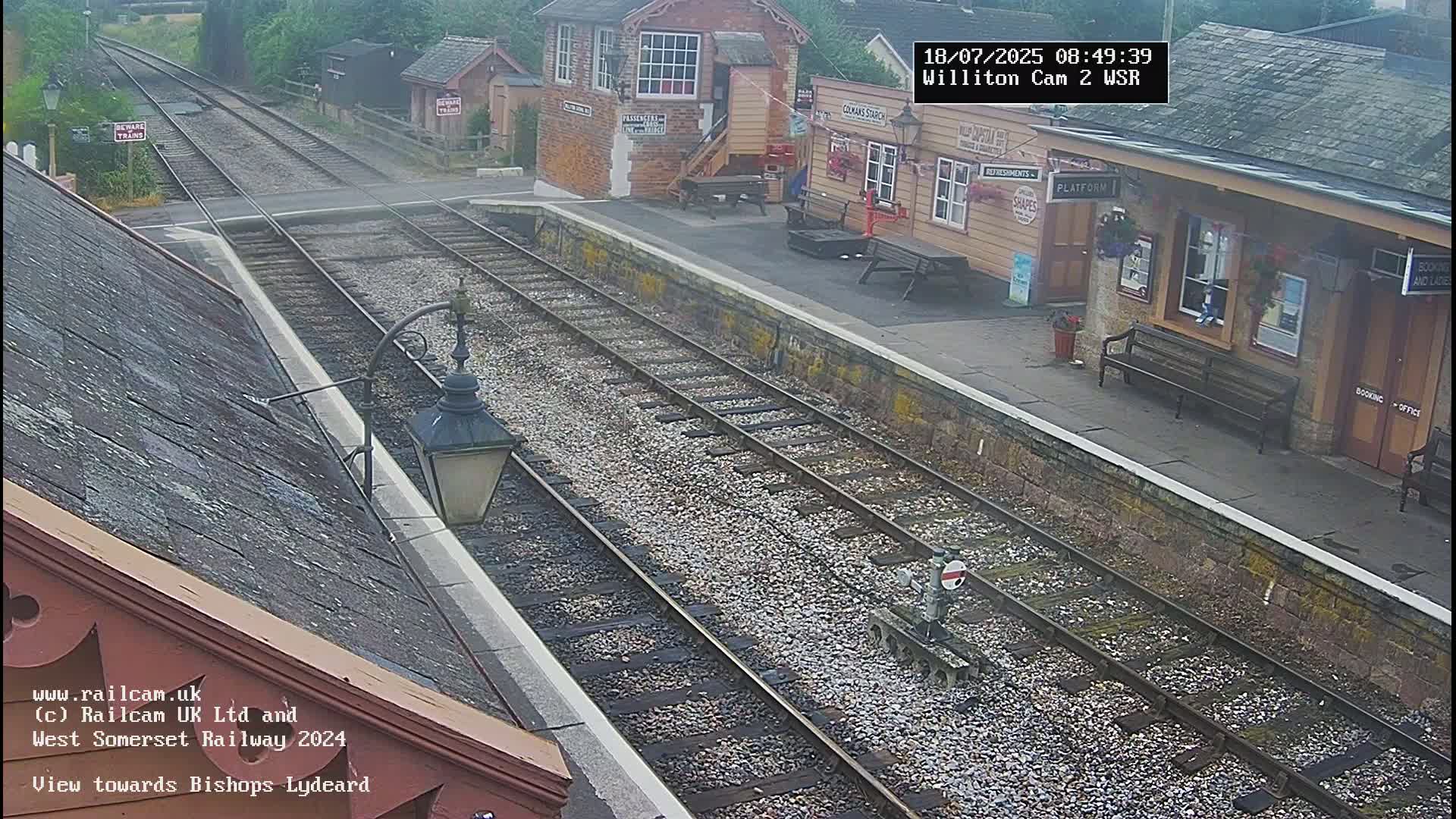 A high-angle view shows a train station on a cloudy day, with train tracks extending into the distance, and a small building with a platform.