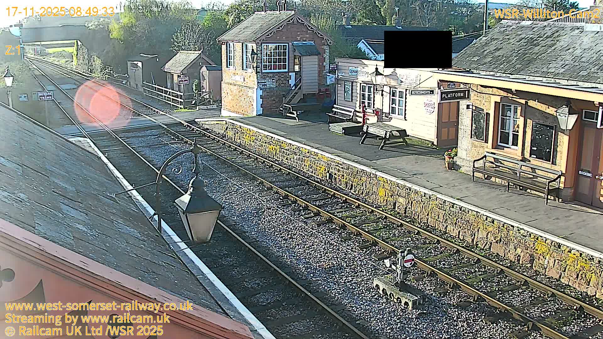 A sunny and clear elevated view captures a vintage rural train station with multiple tracks running alongside a stone-faced platform, featuring traditional station buildings, benches, and old-style lamps.