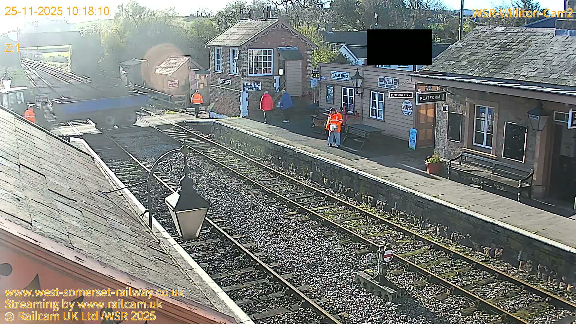 On a bright, sunny day, a train station features a blue maintenance vehicle on the tracks, with several people, some in high-visibility gear, walking on the platforms and near various station buildings.