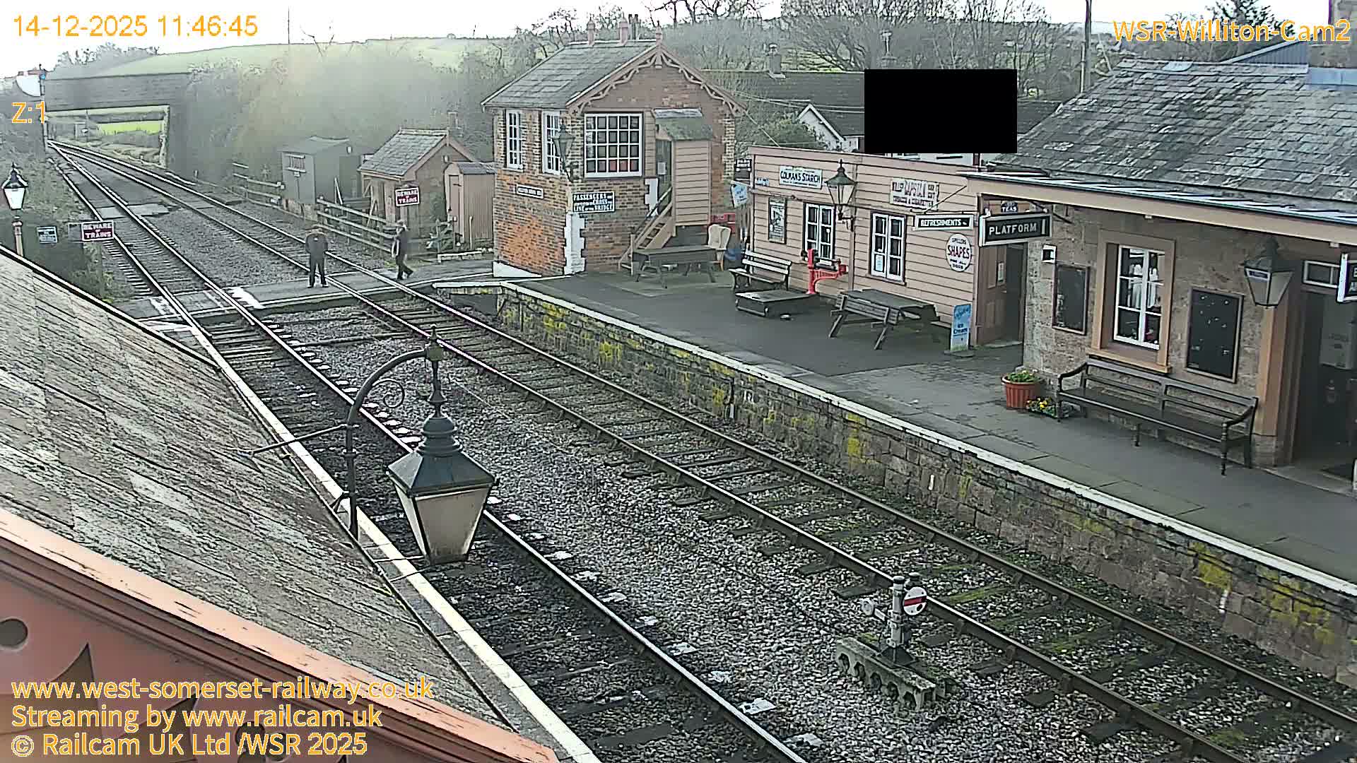 A vintage railway station features multiple gravel tracks leading to an overhead bridge, alongside a brick signal box and a platform with several wooden station buildings, all under an overcast sky.