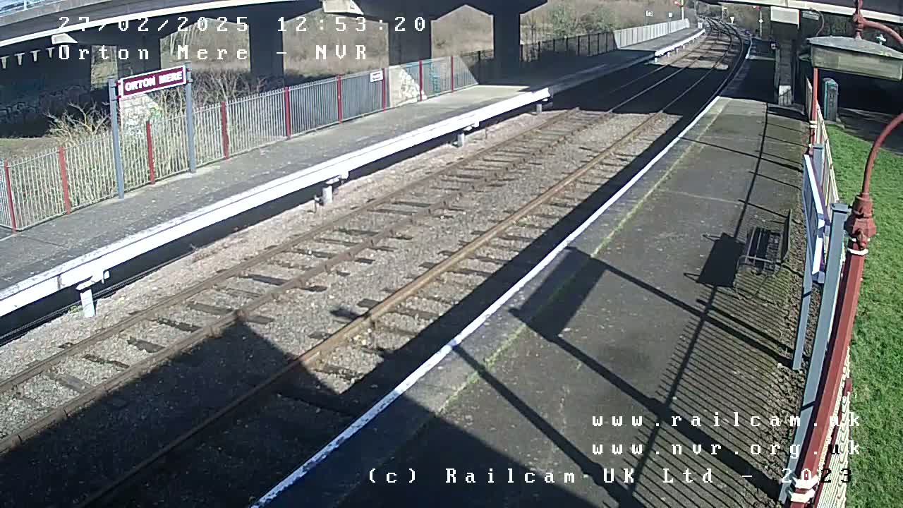 A sunny view of a deserted train platform with railway tracks extending into the distance under a bridge.