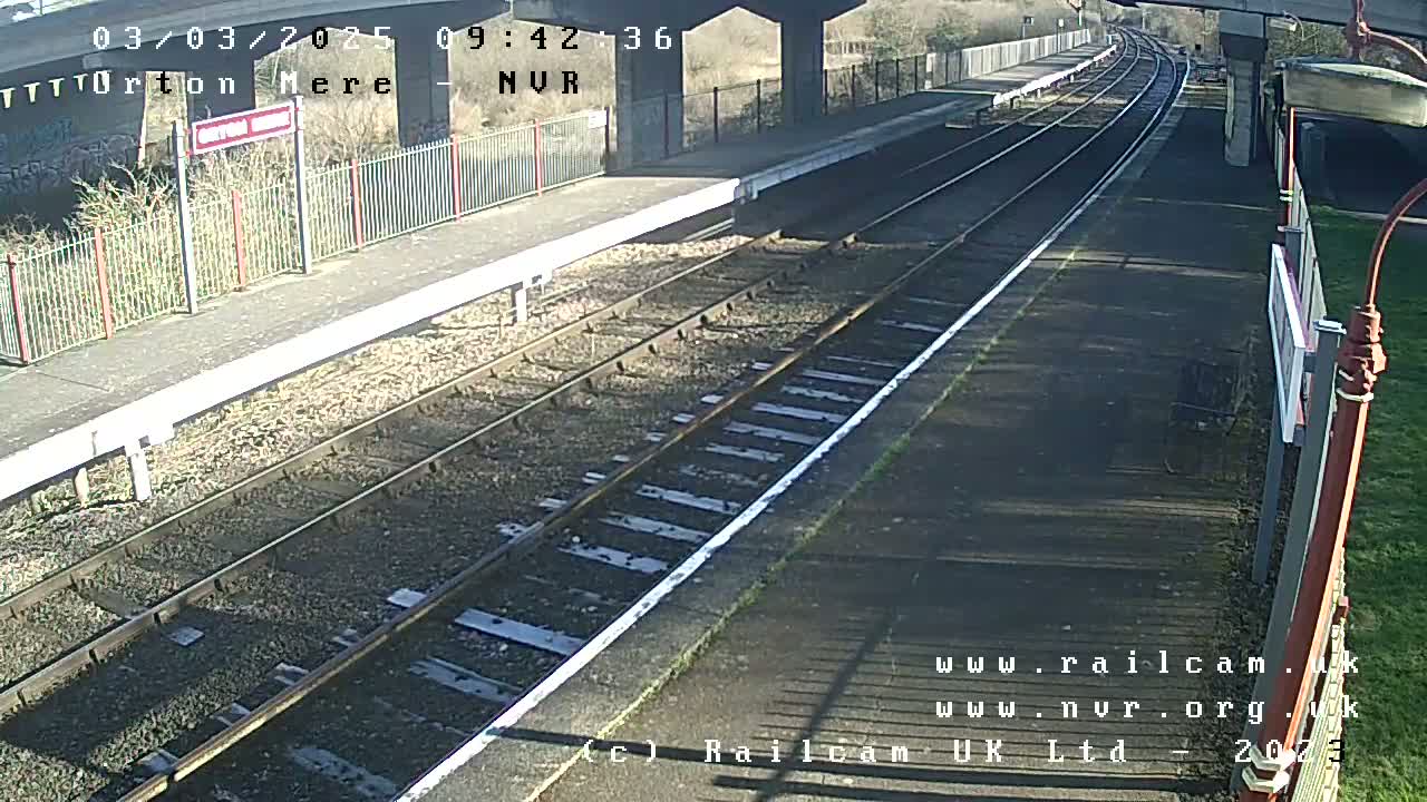 A sunny view of a train station platform with two sets of train tracks extending into the distance.