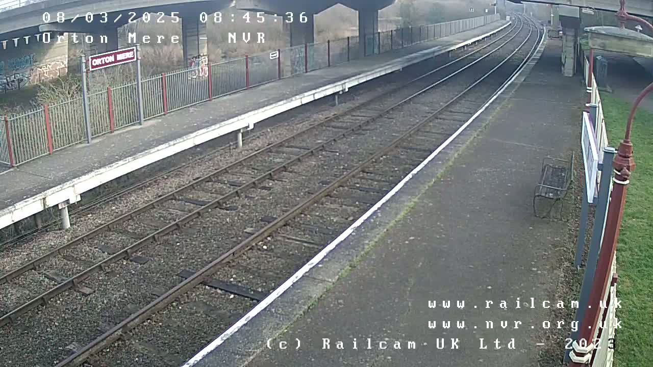 A view of a train platform on a partly cloudy day, showing two sets of train tracks leading to an overpass.