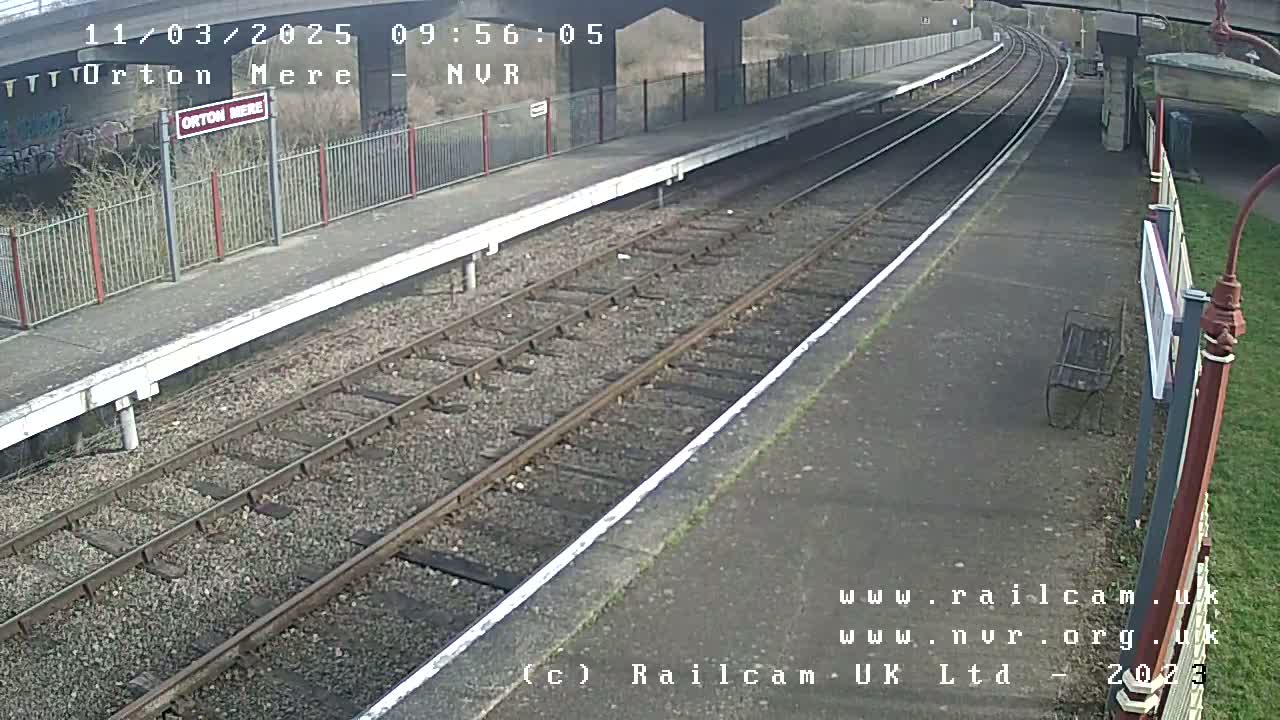 A mostly empty train platform on a sunny day, with train tracks extending into the distance under a bridge.