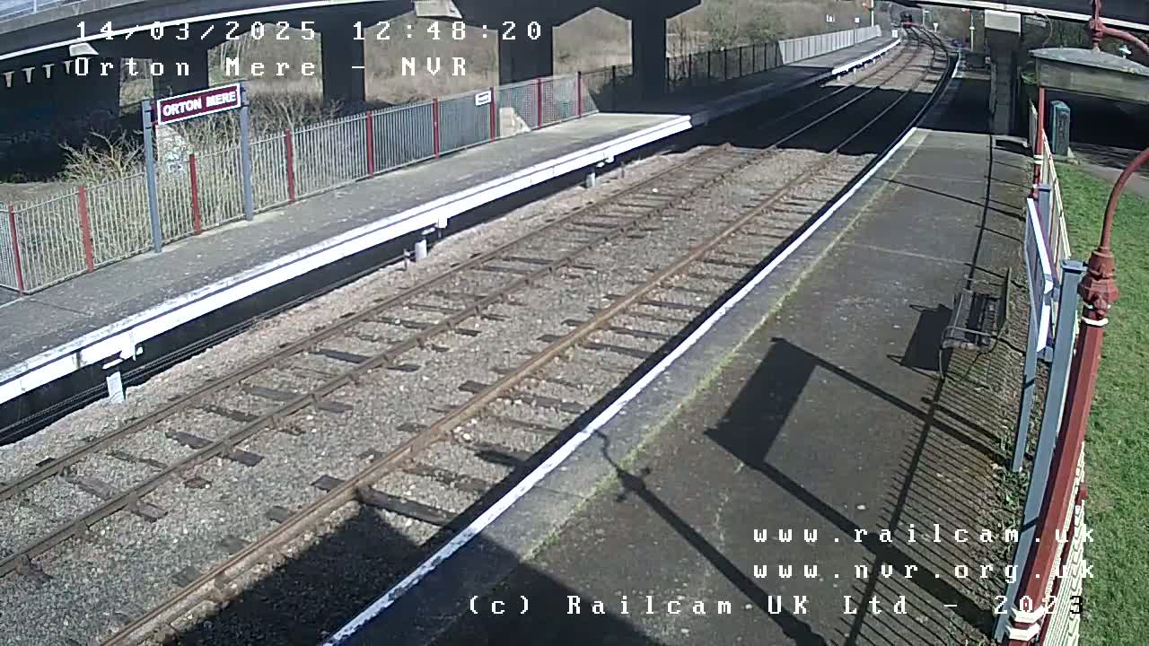 A sunny view of a train station platform with railway tracks extending into the distance, under a bridge.