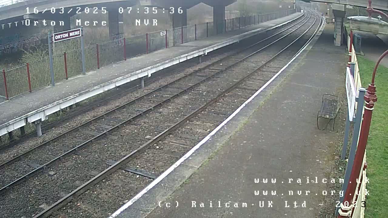 A mostly overcast view of an empty train platform with two sets of train tracks, a bench, and sparse vegetation.