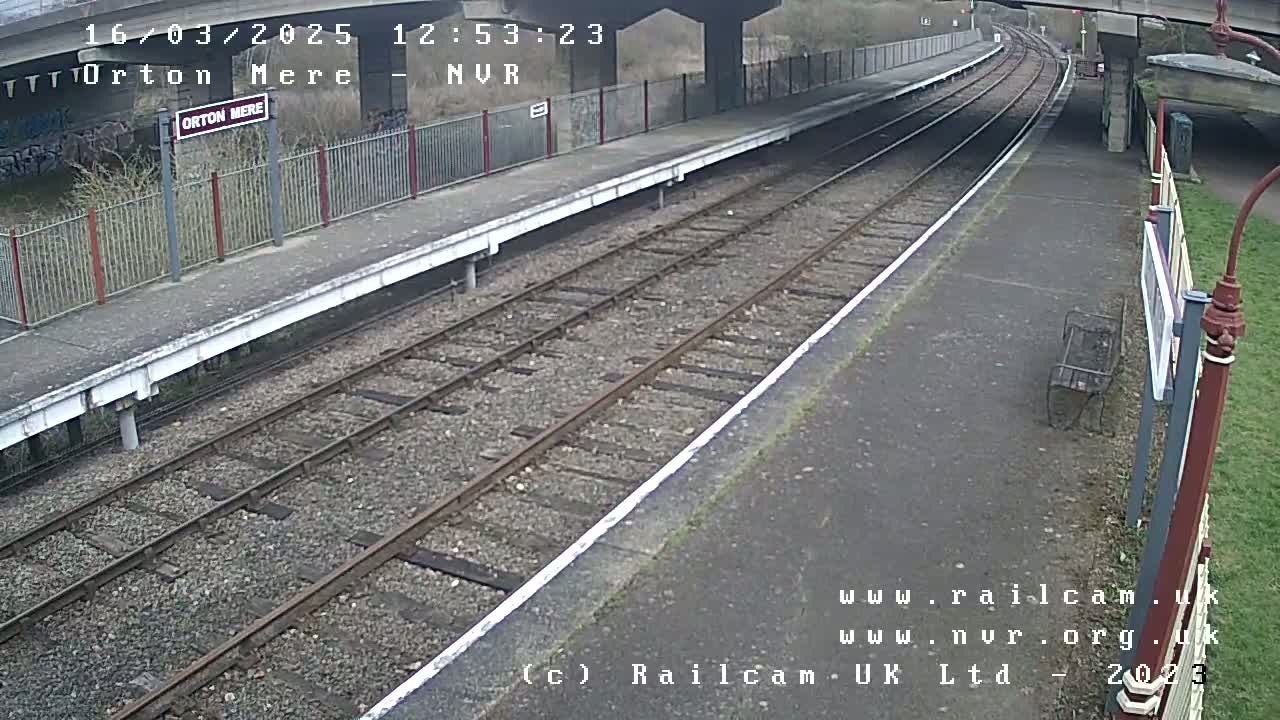 A mostly empty train station platform under a bridge on an overcast day, with train tracks extending into the distance.