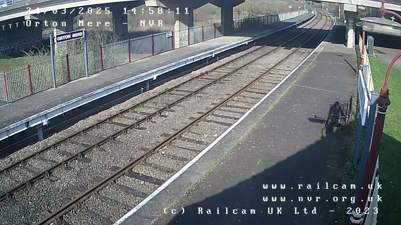 A sunny view of a train station platform with railway tracks extending into the distance under a bridge.