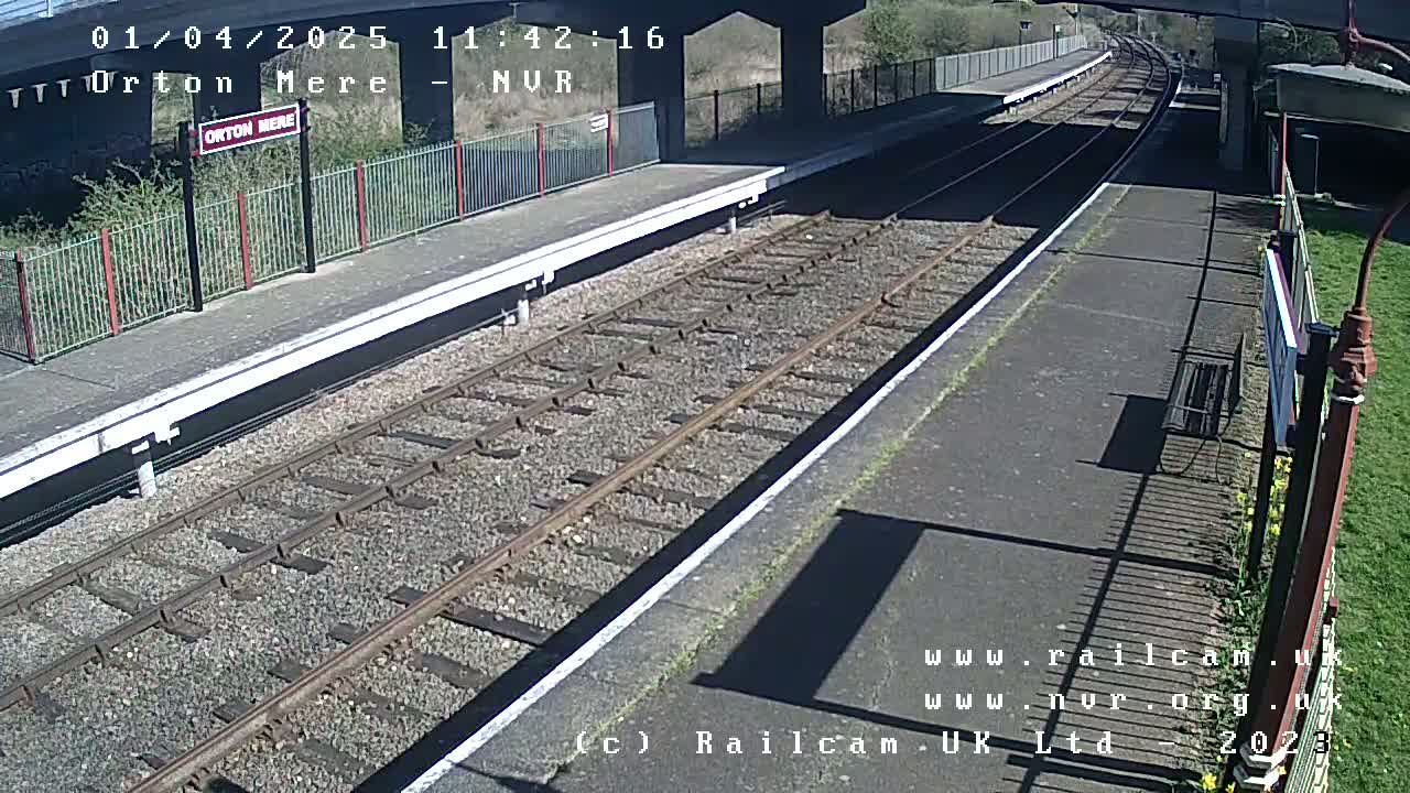 A sunny view of a train station platform with two parallel train tracks extending into the distance under a bridge.
