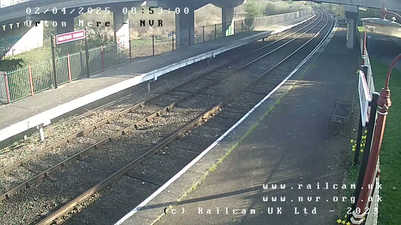 A sunny view of a train station platform with two sets of train tracks and a metal fence.