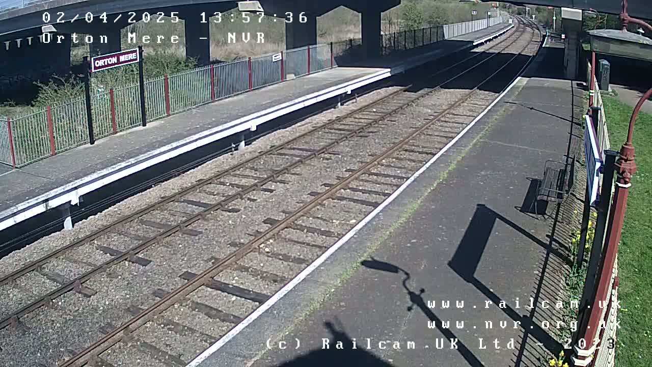 A sunny view of a train platform with railway tracks extending into the distance, under a bridge.