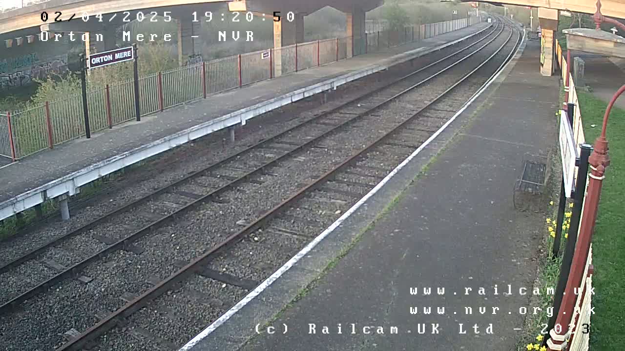 A mostly clear view of a train station platform with two parallel train tracks, under an overcast sky.