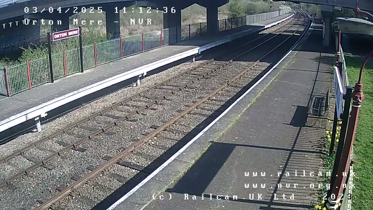 A mostly sunny view of an empty train platform with railway tracks extending into the distance, under a bridge.