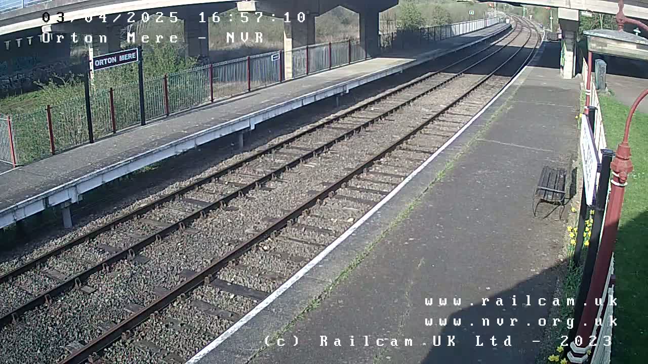 A mostly sunny view of a train station platform with railway tracks extending into the distance under a bridge.