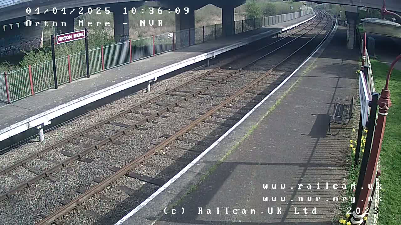 A mostly sunny view of a deserted train platform with two sets of railway tracks and a bench.