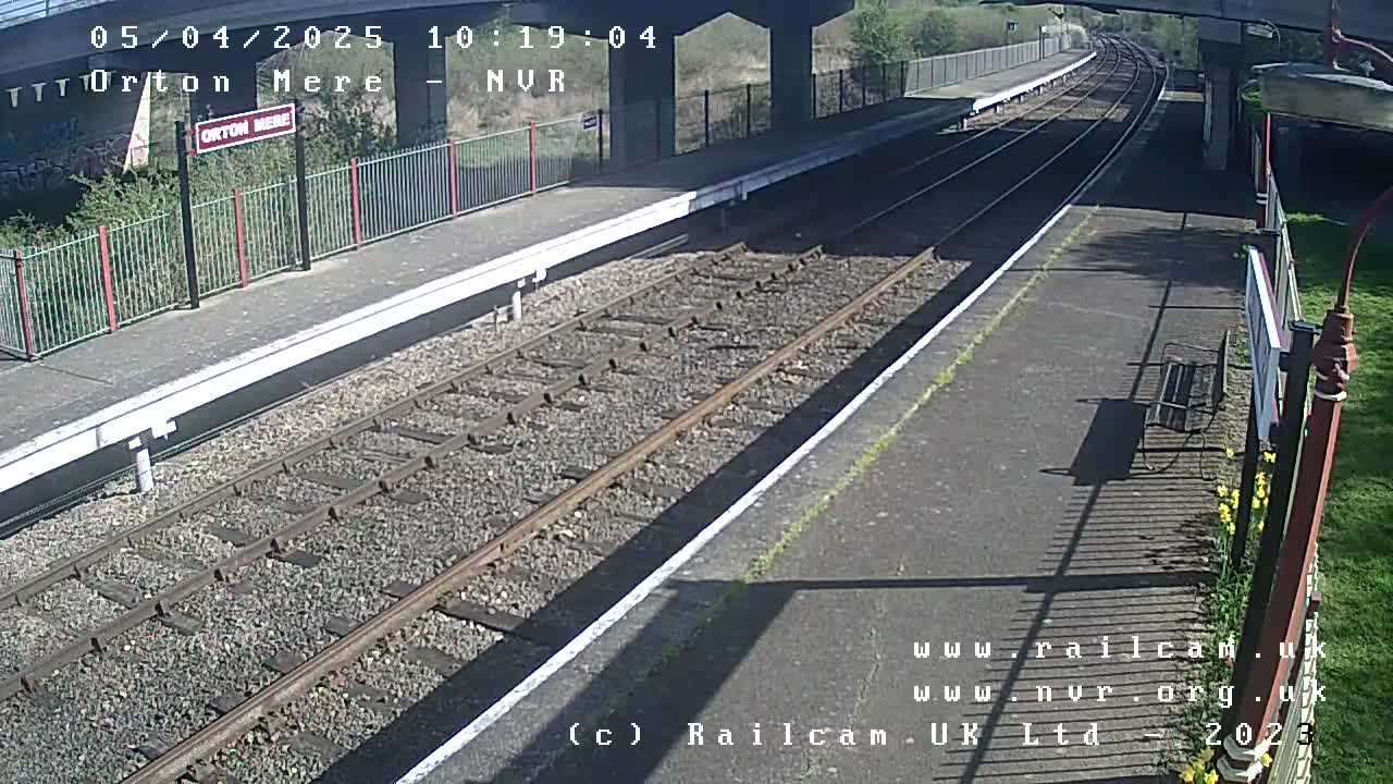 A sunny view of a train station platform with two sets of railway tracks extending into the distance, under a bridge.